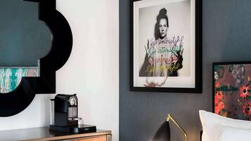 A modern hotel room with a bed, a bedside table with a lamp, a coffee maker, and a framed quote on the wall.