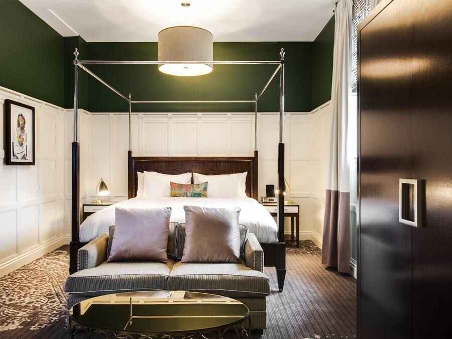 A four-poster bed with a canopy, a bedside table with a lamp, and a small round table in a room with green walls and a patterned carpet.