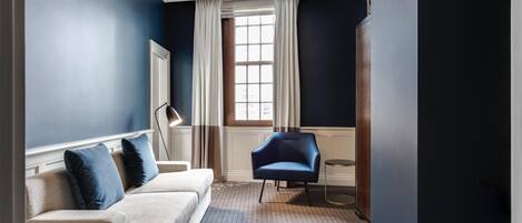 A modern room with a blue accent wall, a white sofa with blue cushions, a blue armchair, a small round table, and a large window with curtains.