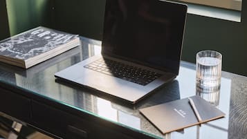 Ein Laptop steht auf einem Glastisch, neben ihm liegen ein Buch und ein Glas Wasser.