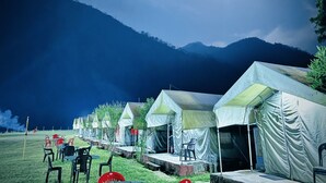 A row of tents with tables and chairs in a grassy field, set against a mountainous backdrop. - Riverstone Camps and Resorts (Yamkeshwar)