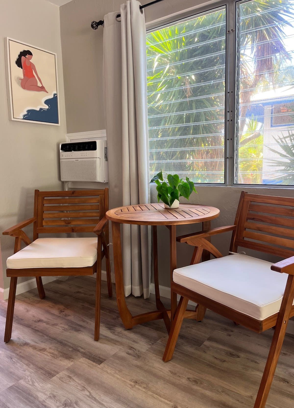 A room with a wooden chair and a table, a window with curtains, and a wall-mounted air conditioner.