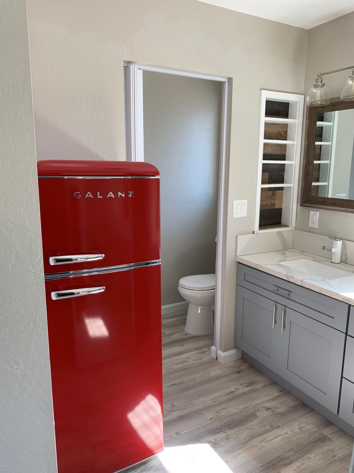 A compact bathroom with a red Galanz refrigerator, a white toilet, a vanity with a mirror, and a wooden floor.