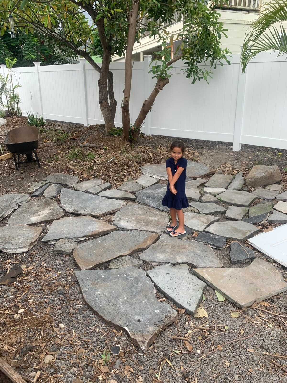 Un enfant se tient sur un chemin de pierres dans un jardin, avec une clôture blanche et des arbres en arrière-plan.