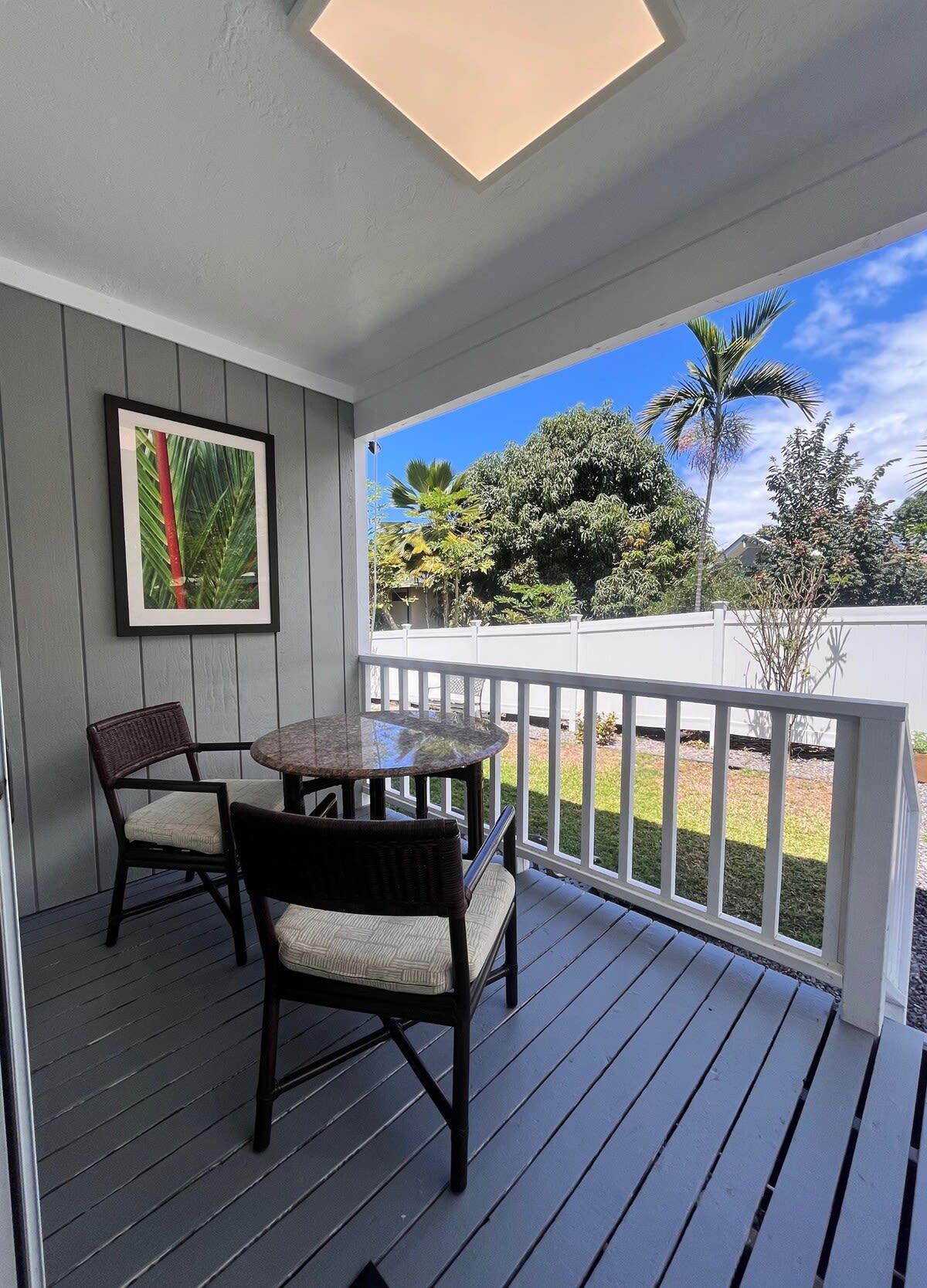 A covered outdoor area with a table and chairs, a framed picture on the wall, and a view of a garden with trees and a white fence.