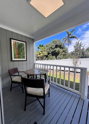 Varanda coberta com mesa e cadeiras, quadro na parede e vista para um jardim com árvores e palmeiras.