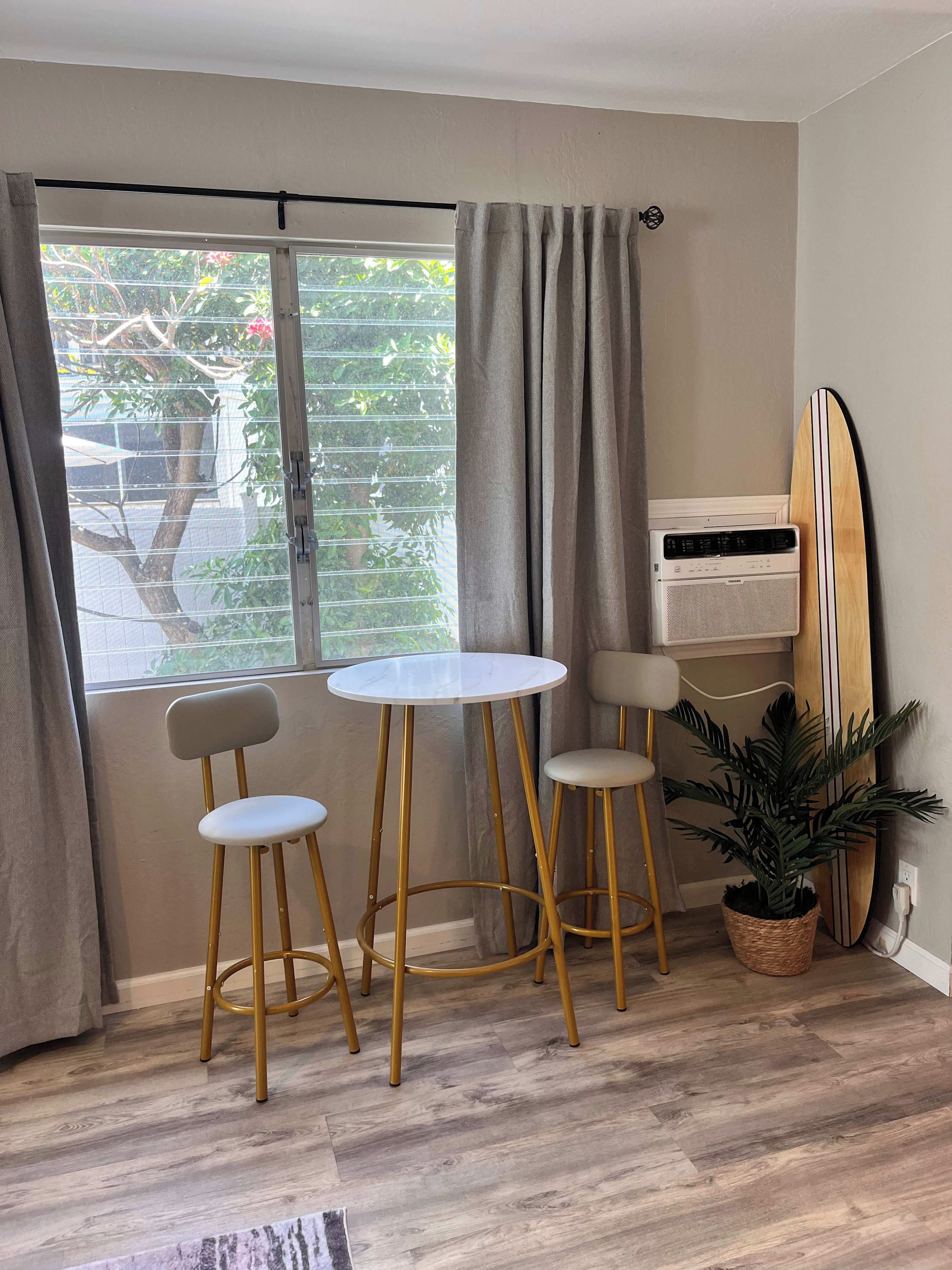 Een kamer met een raam, twee stoelen, een klein tafeltje, een surfboard en een potplant.