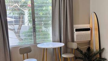 Een kamer met een raam, twee stoelen, een klein tafeltje, een surfboard en een potplant.
