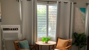 Una habitación con una mesa redonda de madera, dos sillas de madera con cojines, una planta en maceta y un cuadro con motivos playeros en la pared.