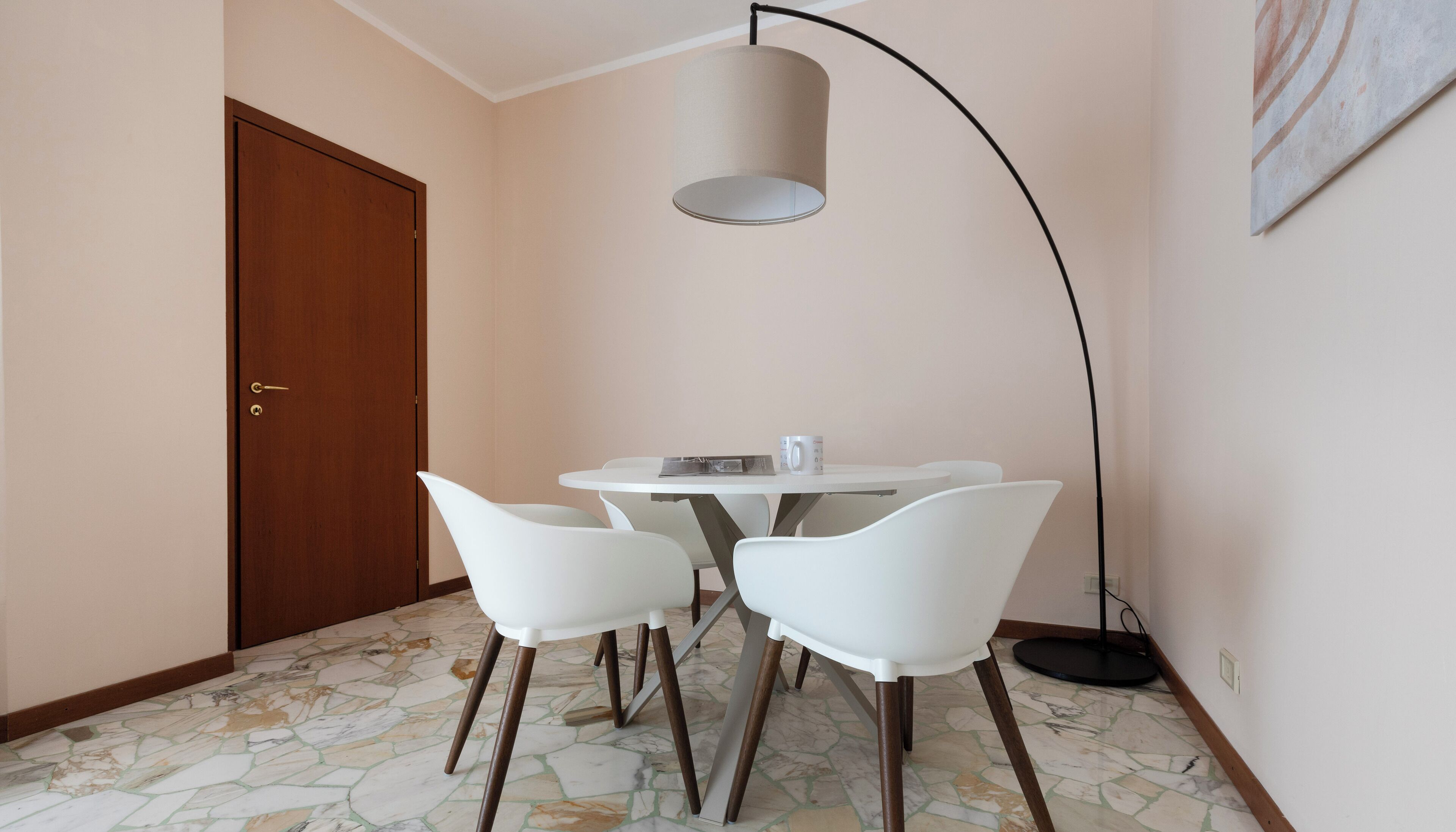 A room with a wooden door, a white table with two chairs, a floor lamp, and a tiled floor.