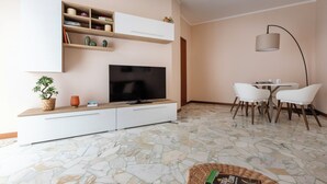 A modern living room with a flat-screen TV, white cabinets, a dining area with a table and chairs, and a patterned floor. - Italianway - Campania 26 A (Milan)