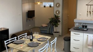 Une cuisine moderne avec des meubles blancs, un coin repas comprenant une table en bois et des chaises blanches, une plante en pot et un téléviseur fixé au mur.