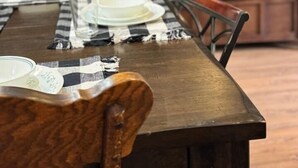 Ein Essbereich mit einem Holztisch, gedeckt für zwei Personen, einem an der Wand montierten Fernseher und einem Fenster mit Jalousien.
