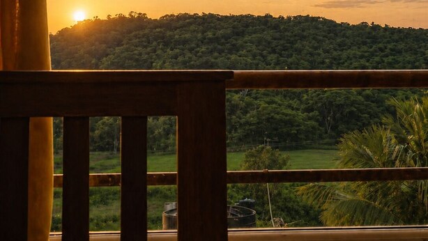 Une vue sur le coucher de soleil depuis un balcon doté d’une balustrade en bois, avec une colline boisée en arrière-plan et un ciel dégagé.