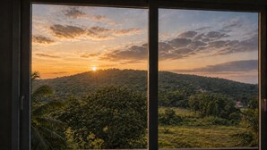 Une vue à travers une fenêtre sur un paysage comprenant une colline, des arbres et une maison au loin.