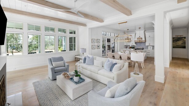 A spacious living room with a large white sofa, a grey armchair, and a coffee table. There is a dining area with a white table and chairs, and a kitchen with white cabinets and a chandelier. - Poolside Privacy & Dining Under the Stars (Bluffton)
