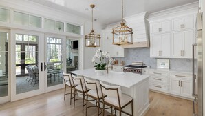 A spacious kitchen with white cabinetry, a large island with bar stools, and a view through glass doors to an outdoor patio. - Poolside Privacy & Dining Under the Stars (Bluffton)