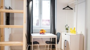 Une pièce de taille modeste, avec une table blanche, deux chaises, un radiateur, une fenêtre avec des rideaux, une armoire blanche et un porte-vêtements suspendu.