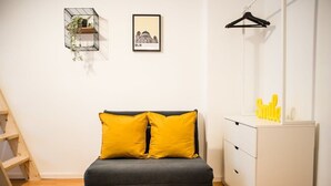 A modern bedroom with a grey sofa bed, two yellow pillows, a white dresser, a black hanger, and a framed picture on the wall. - Cosy Box @ Boxhagener Platz (Berlin)