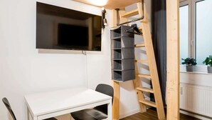 A room with a white desk, black chairs, a TV mounted on the wall, a wooden ladder, and a window with a view of the outside. - Cosy Box at Boxhagener Platz (Berlin)
