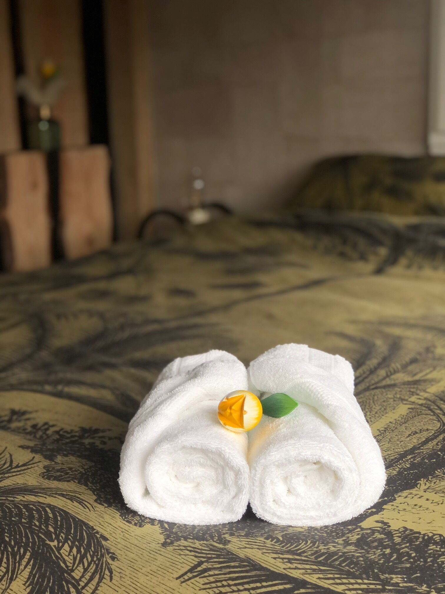 A neatly folded towel with a decorative item on a patterned bedspread in a room with a wooden headboard and a vase of flowers in the background.