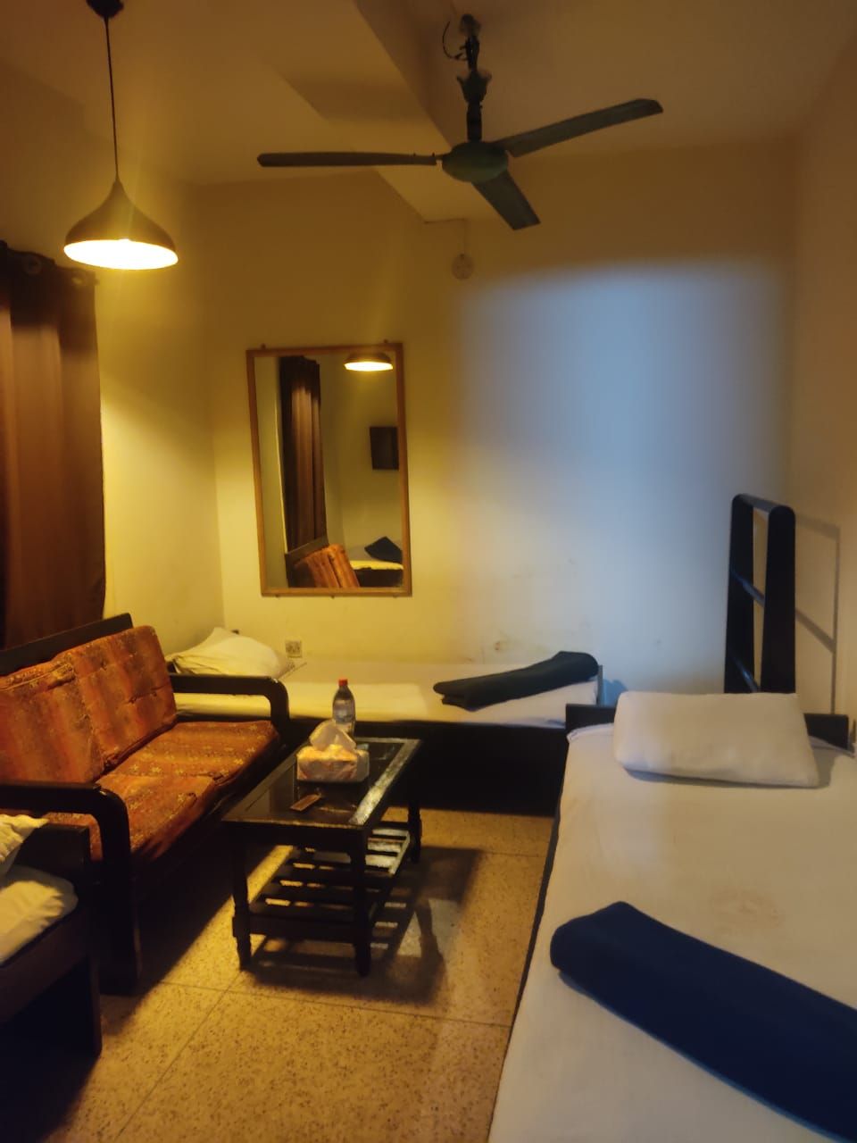 A hotel room with a sofa, a small table, a bed, a ceiling fan, a mirror, and a TV.