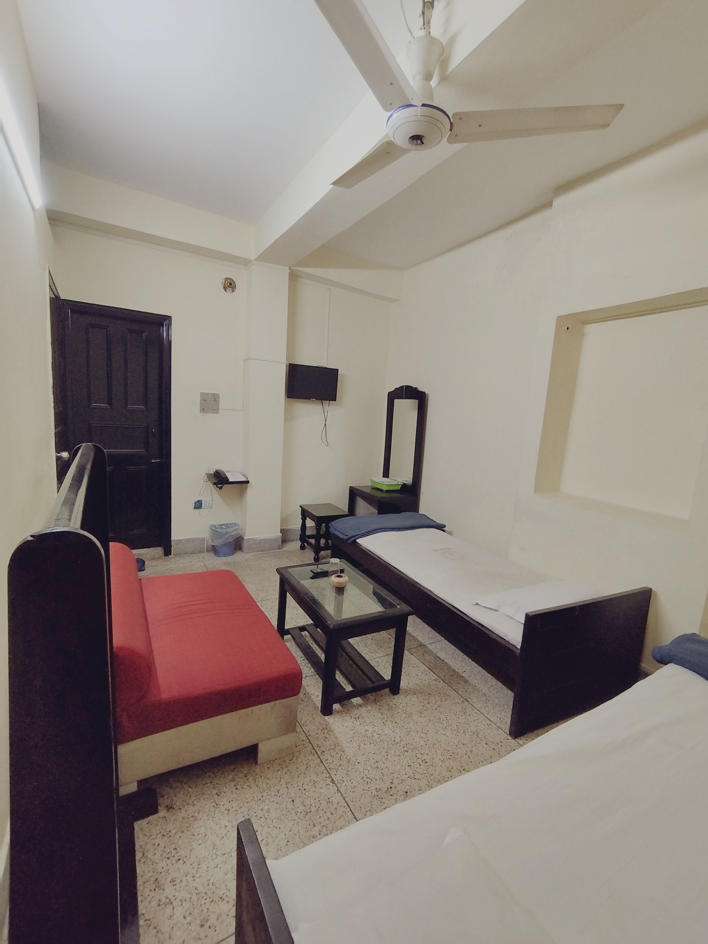 Una stanza con due letti singoli, un divano rosso, un tavolino, una ventola a soffitto, una TV e una porta.