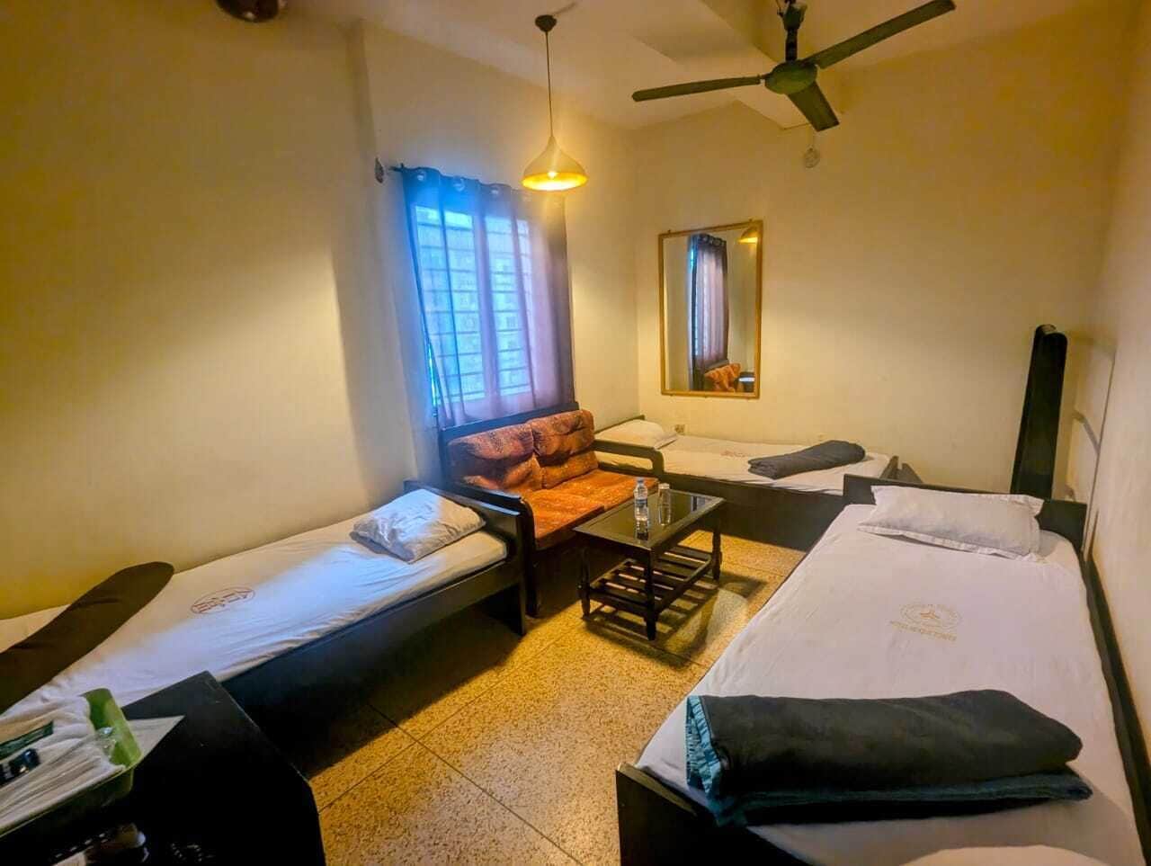 A small, well-lit room with two single beds, a sofa, a table, and a ceiling fan.