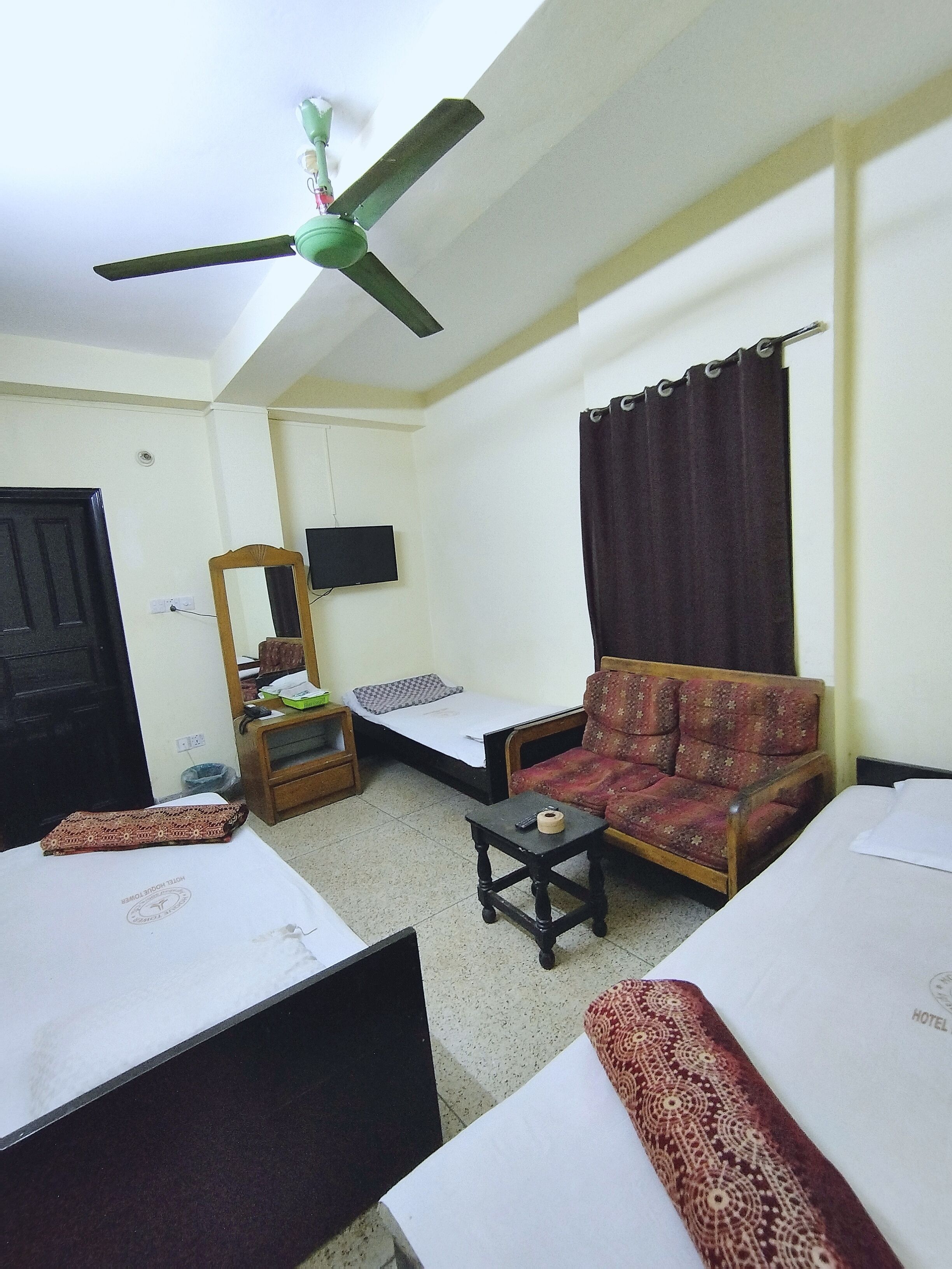 Habitación de hotel con dos camas individuales, un sofá, una mesita, ventilador de techo, televisor y ventana con cortinas.