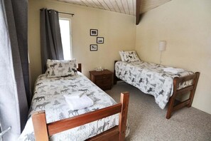 Ein Zimmer mit zwei Holzbettgestellen, Bettwäsche mit Tiermotiven, einem kleinen Nachttisch, einer Wandlampe und einem Fenster mit Vorhängen.