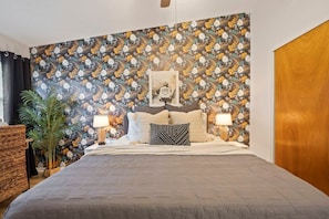 A bedroom with a patterned wallpaper, a bed with multiple pillows, two bedside lamps, and a framed picture on the wall.