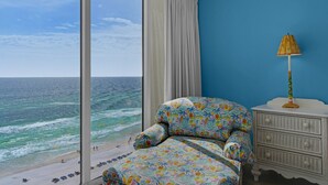 Een kamer met een groot raam met uitzicht op de oceaan, een stoel met bloemenprint, een witte dressoir en een lamp.