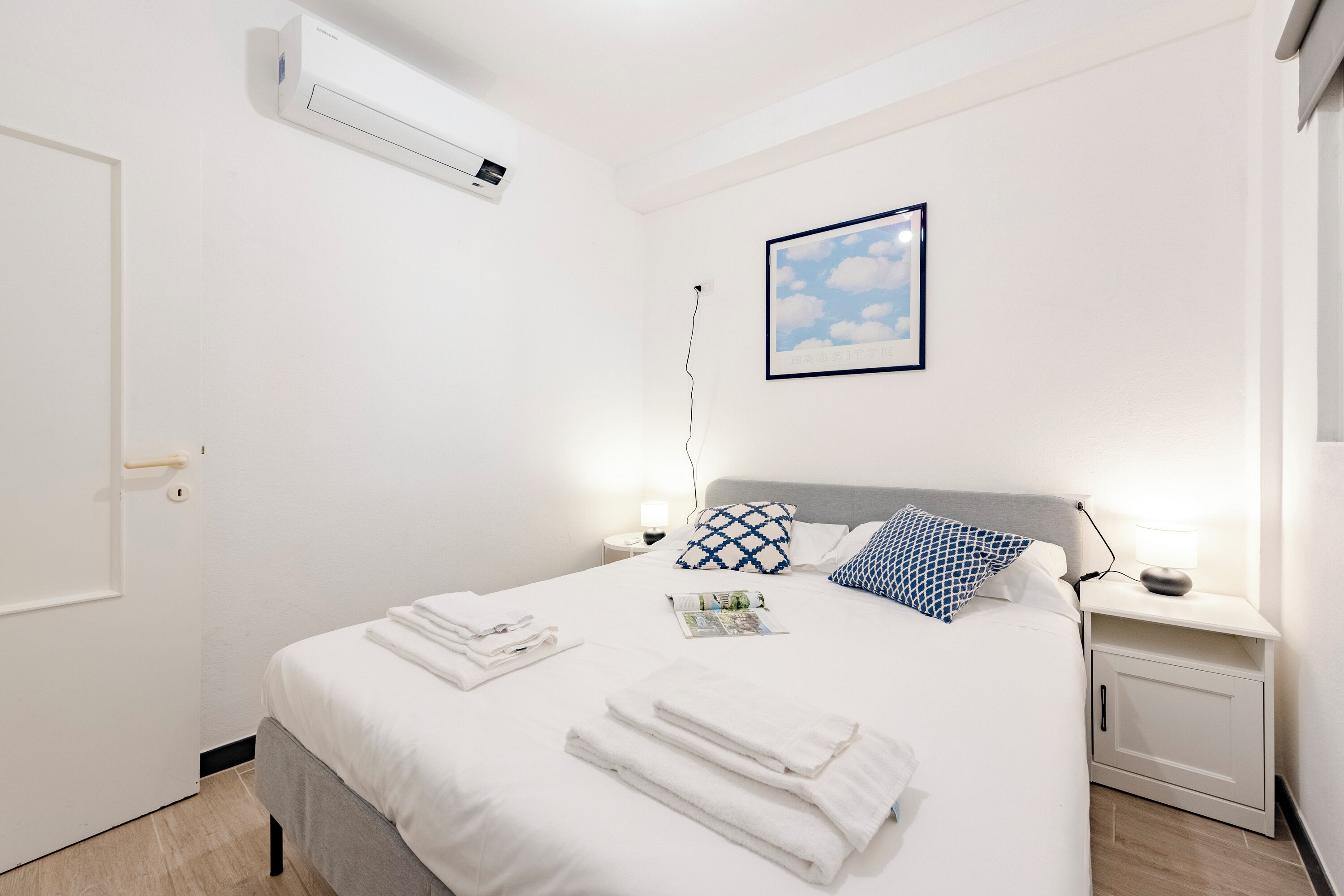 Ein ordentlich bezogenes Bett mit weißen Leinen und Handtüchern, eine Nachttischlampe, ein gerahmtes Bild von Wolken und eine weiße Klimaanlage an der Wand.
