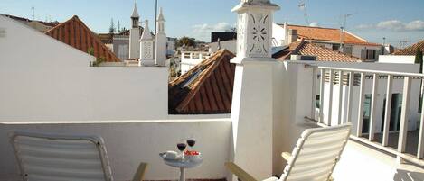 A terrace with two white chairs, a small table with a wine glass and a bowl of fruit, and a white clock tower in the background.