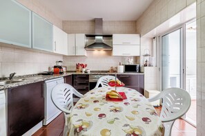Une cuisine avec des meubles blancs, une hotte de cuisinière en acier inoxydable et une table à manger recouverte d’une nappe à motifs de fruits.