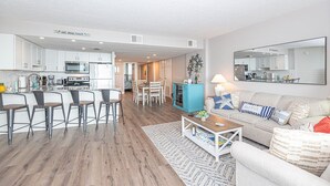 Eine moderne Küche mit weißen Schränken, ein Essbereich mit Tisch und Stühlen, ein Wohnzimmer mit Sofa und Couchtisch sowie ein großer Spiegel an der Wand.