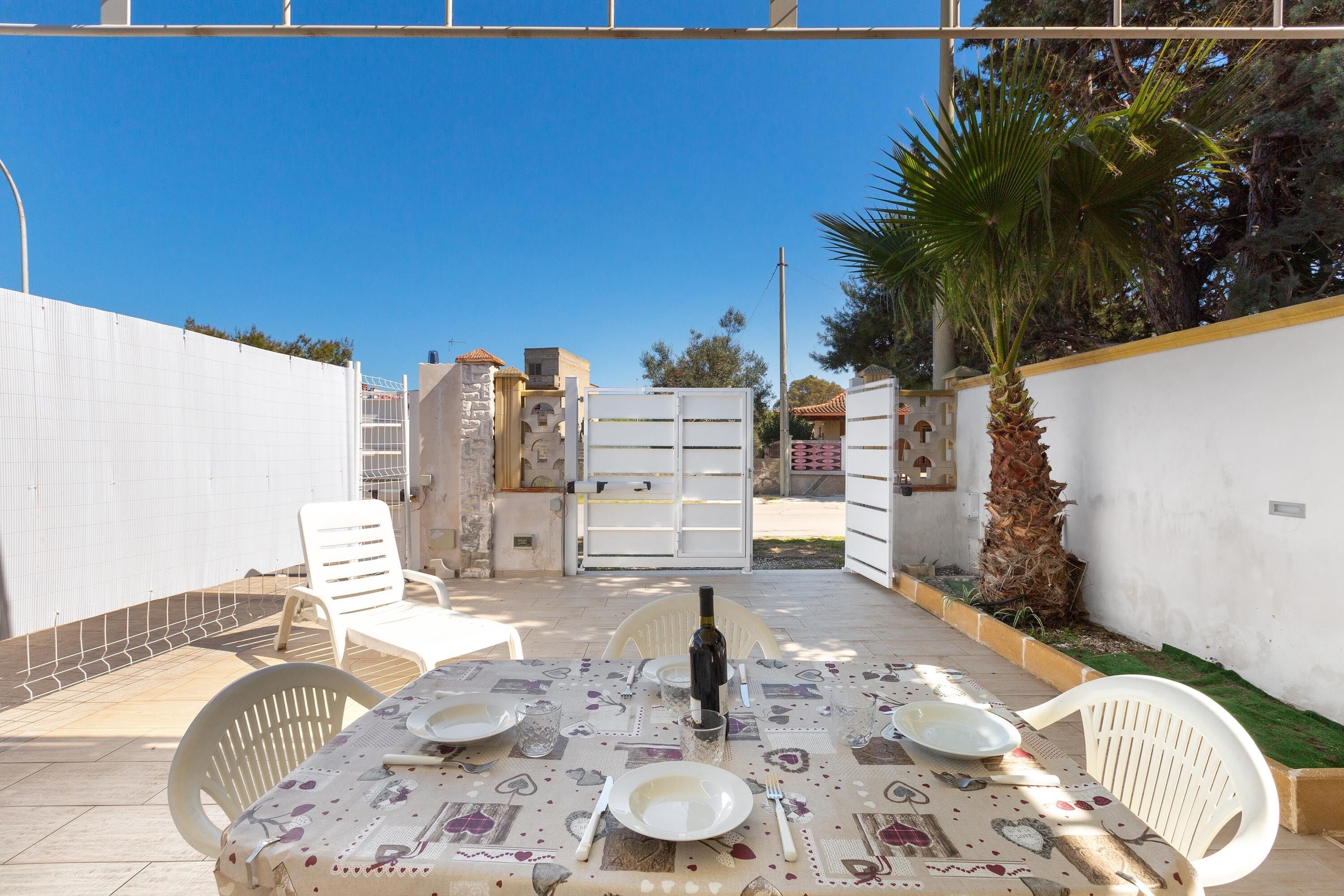 Una terrazza con tavolo apparecchiato per mangiare, circondata da sedie bianche, una bottiglia di vino e una ciotola.