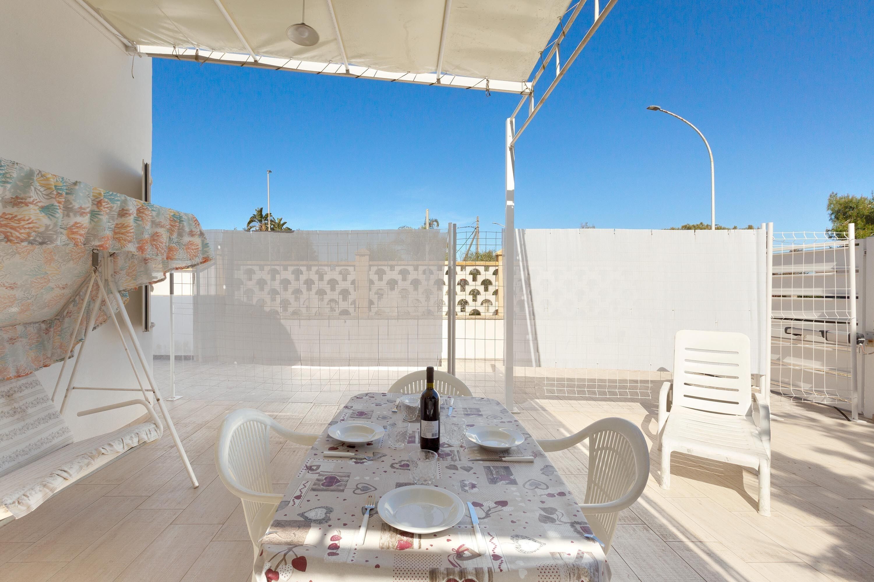 Un patio al aire libre con una mesa lista para dos personas, una botella de vino y un mantel estampado.