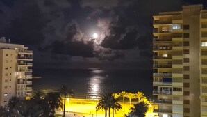 A night view of a coastal area with a building on the left and a brightly lit area on the right.