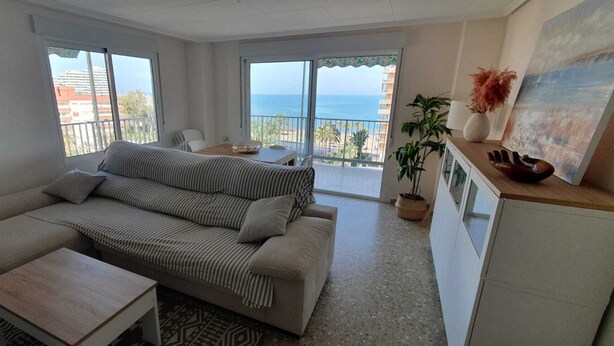 A living room with a sofa, a small table, and a balcony with a view of the sea.