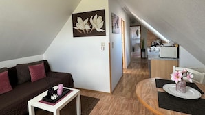 Un appartement moderne de style loft, doté d’un plafond en pente, d’un espace de vie avec un canapé et une table basse, d’un coin repas avec une table ronde et d’une petite cuisine en arrière-plan.