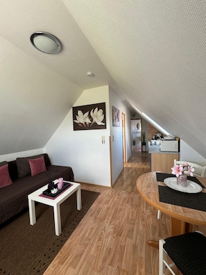 Een modern loft appartement met schuin plafond, een zithoek met bank en salontafel, een eethoek met ronde tafel, en een kitchenette op de achtergrond.