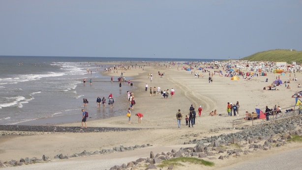 Une plage bondée, où l’on voit des gens se promener et rester debout, des tentes et une côte rocheuse.