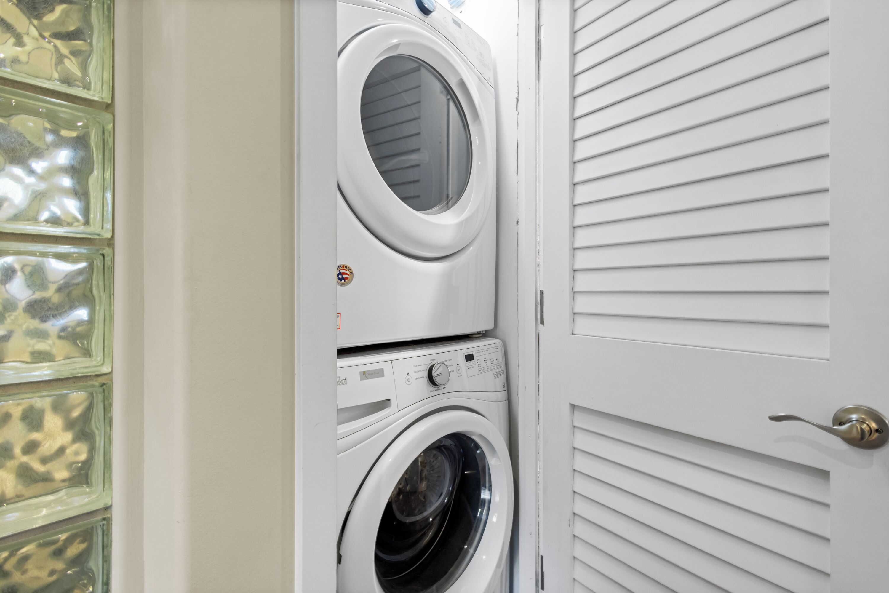 Un lavadero con dos lavadoras superpuestas y una puerta con persianas horizontales.
