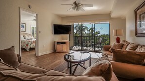 A living room with a brown leather sofa, a glass coffee table, a ceiling fan, a flat-screen TV, and a balcony with a view of palm trees. - 2B/2B ISLAND VACATION CONDO. RESORT POOL! WALK TO BEACH! BEST PRICE! (Fort Myers Beach)