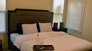 A neatly made bed with a dark headboard, a tray with a bottle and glasses, and a nightstand with a candle and a small plant. - Garden Cottage in the city. (West Monroe)