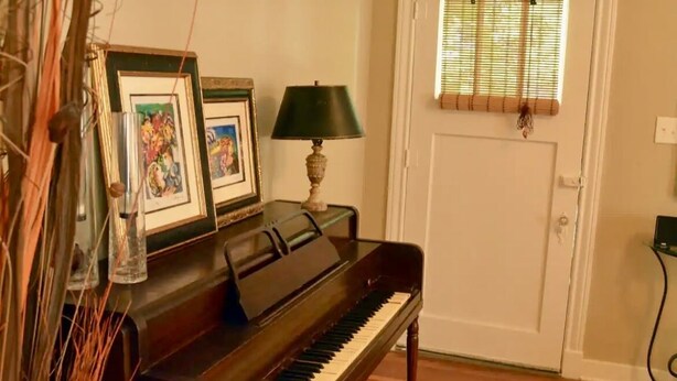A piano in a room with a door, a window with blinds, and framed pictures on the wall. - Garden Cottage in the city. (West Monroe)