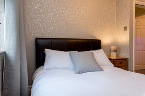 A neatly made bed with white linens and two pillows, a wooden nightstand with a lamp, and a textured wall. - Cattle Market View Apartment (Boston)