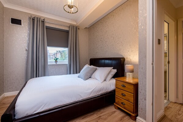 A bedroom with a bed, a nightstand, a lamp, and a window with curtains. - Cattle Market View Apartment (Boston)