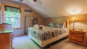 A cozy bedroom with a bed, a bedside table, a lamp, a hanging rack, and a window with a view of greenery.